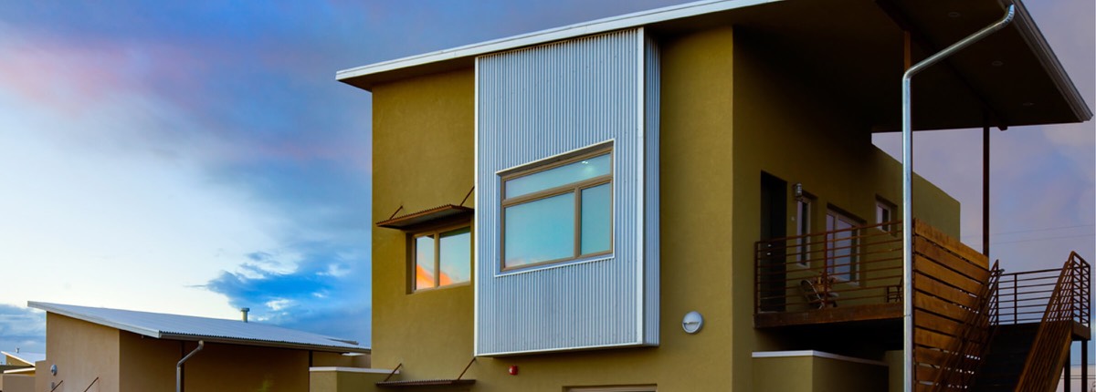 Evening panorama of an olive stucco apartment home with a corrugated steel window bay at La Pradera in Hobbs